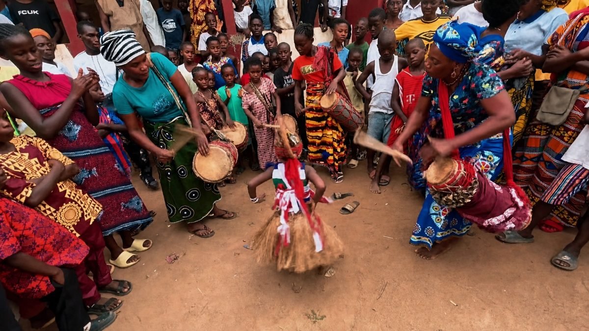 Traditional Drumming and Masked Dance Women playing traditional drums as a masked dancer in a raffia skirt performs at a Liberian community celebration